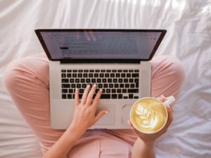 Woman working from home, sitting on a bed with a laptop and coffee
